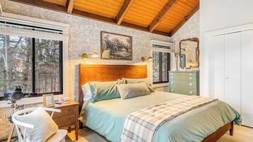 A bedroom with a wooden headboard, a bed with a plaid blanket, a nightstand with a lamp, a dresser, and a window with a view of trees.
