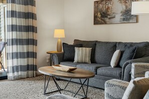 A living room with a grey sofa, a round coffee table with a wooden bowl, a lamp, and a painting of a forest scene. - Enjoy the Indoor Pool, Hot Tub and Sauna! Free Daily Attraction Tickets (Branson)