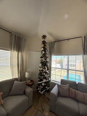 Ein Wohnzimmer mit Weihnachtsbaum, zwei grauen Sofas, einer Lampe und einem Fenster mit Blick auf einen Pool.