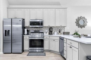Una cucina moderna con mobili bianchi, elettrodomestici in acciaio inossidabile e una parete di fondo rivestita con piastrelle a motivi a spina di pesce.