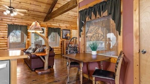 Interior de una cabaña de madera con una mesa de comedor, sillas, un sofá, un ventilador de techo y una ventana con persianas.