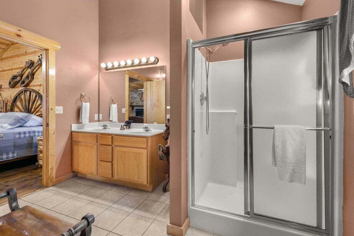 A bathroom with a shower, a vanity with a mirror, and a bed in the adjacent room.