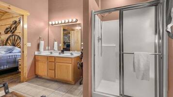 A bathroom with a shower, a vanity with a mirror, and a bed in the adjacent room.