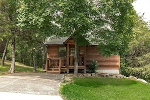 A log cabin with a porch, surrounded by trees and a grassy area.