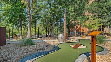 A miniature golf course with a winding path, wooden structures, and a backdrop of trees and buildings.