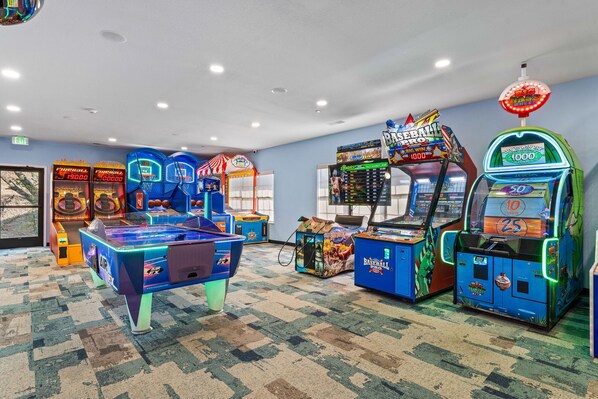 Habitación con máquinas de videojuegos arcade, incluyendo baloncesto y béisbol, piso alfombrado e iluminación empotrada en el techo.