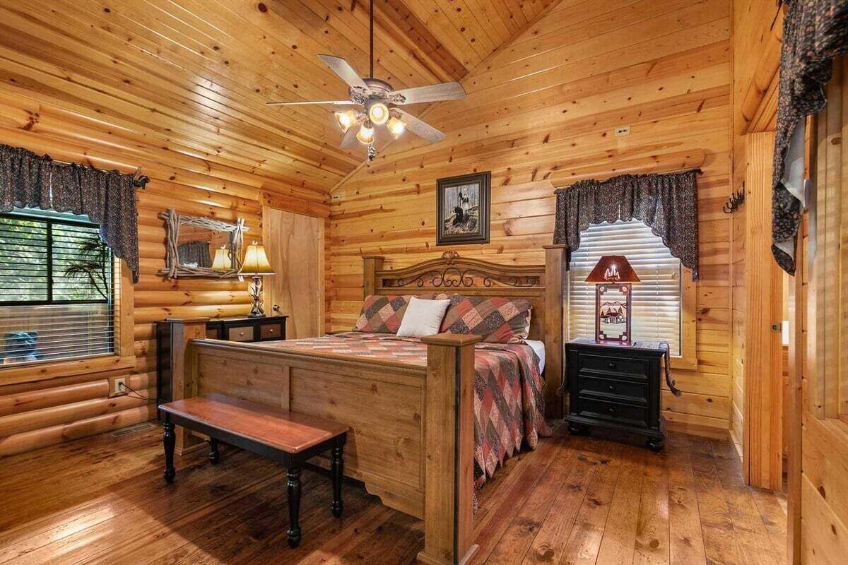 A log cabin bedroom with a wooden bed, a bench, a nightstand, and a ceiling fan.