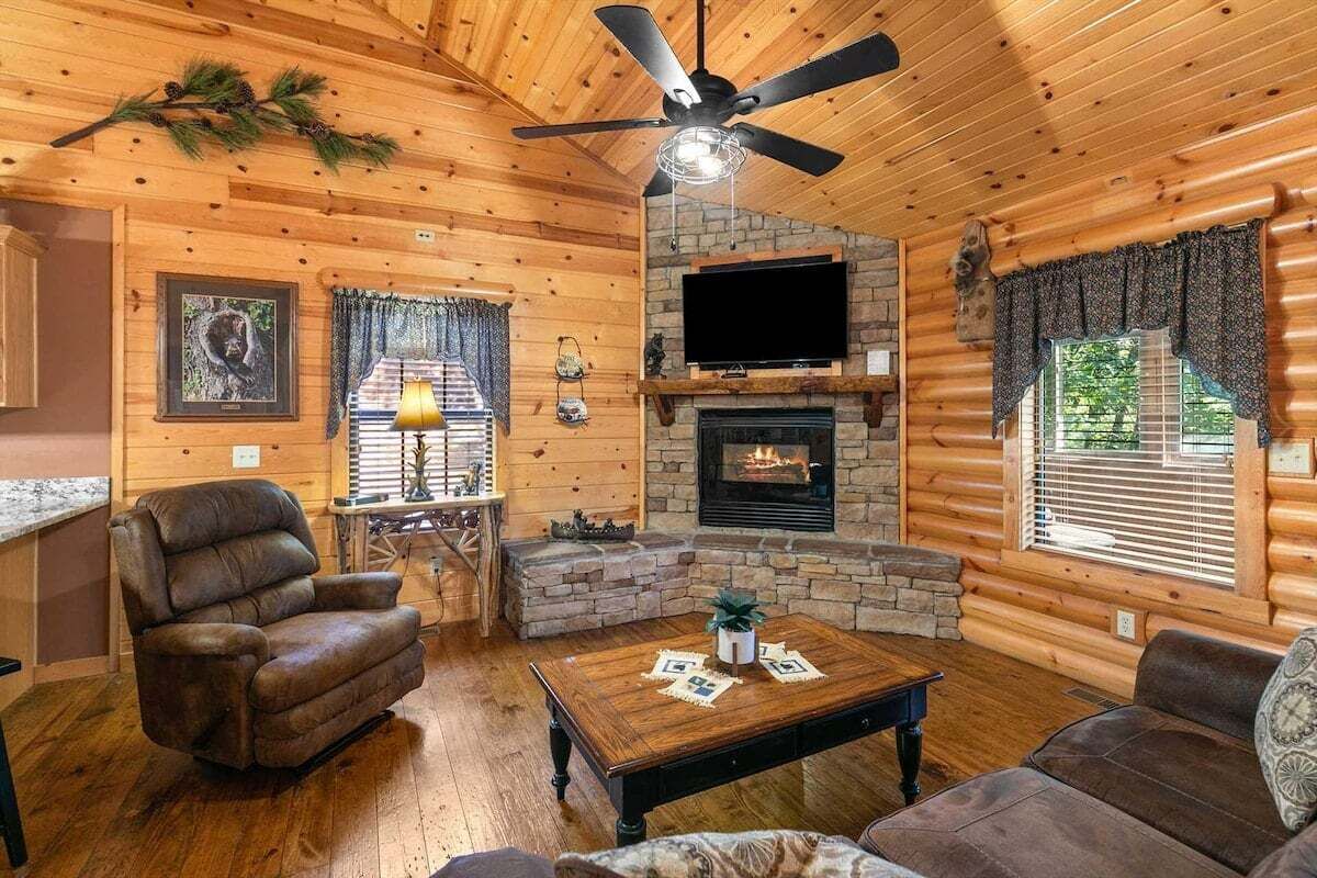 A log cabin interior with a fireplace, a ceiling fan, and a television mounted above it.