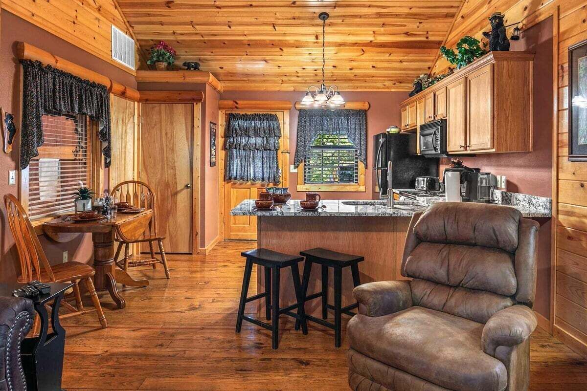 A cabin interior with a wooden ceiling, a dining area with a table and chairs, a bar area with stools, and a kitchen with cabinets and appliances.