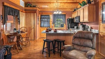 A cabin interior with a wooden ceiling, a dining area with a table and chairs, a bar area with stools, and a kitchen with cabinets and appliances.