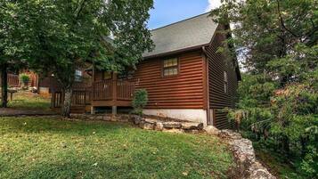 A log cabin with a deck, surrounded by trees and a stone wall.