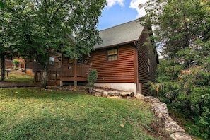 A log cabin with a deck, surrounded by trees and a stone wall.