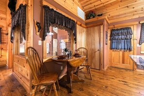 A wooden interior with a dining area featuring a table and chairs, a window with blinds, and a door with a curtain.