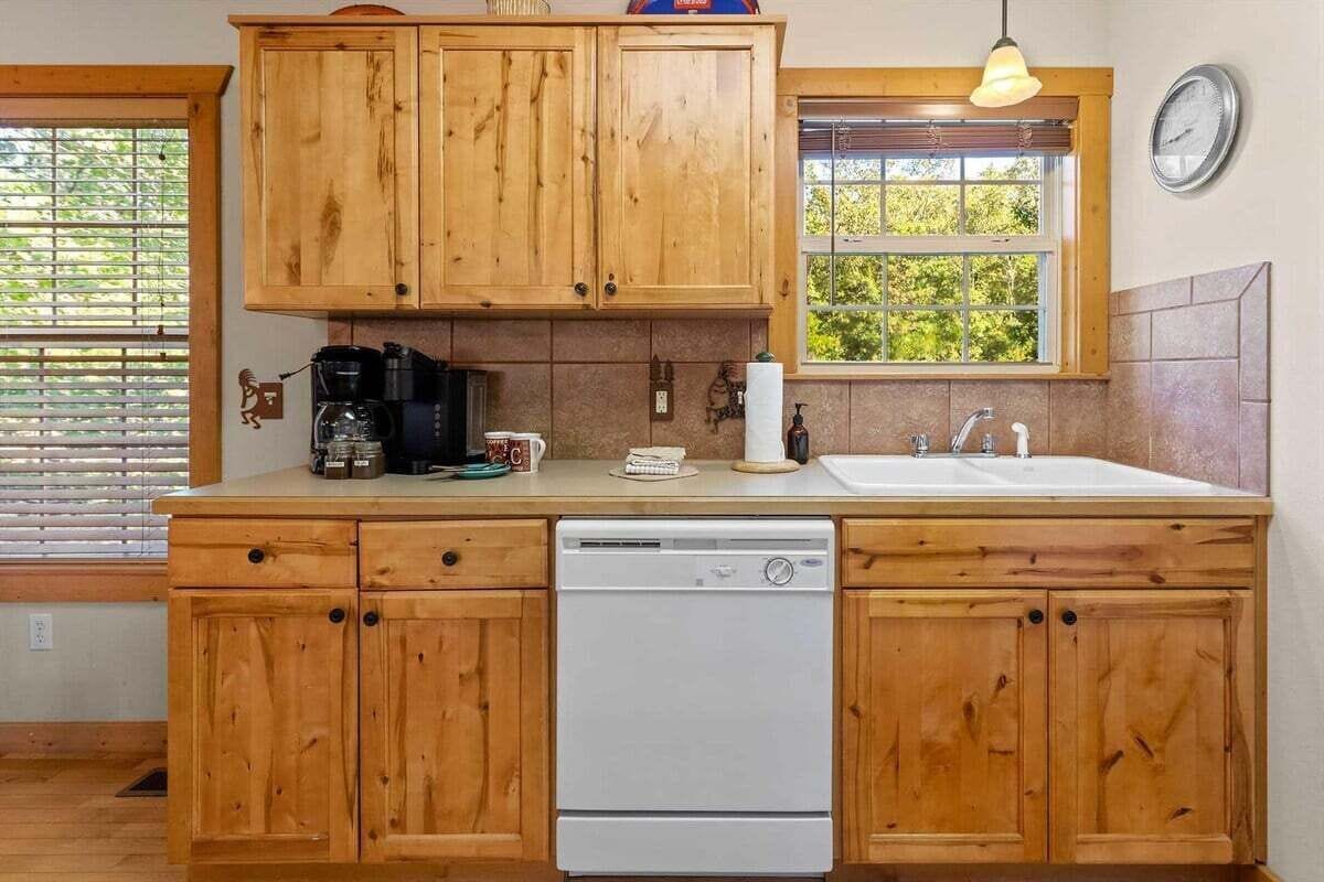 A kitchen with wooden cabinets, a dishwasher, a coffee maker, and a window with blinds.