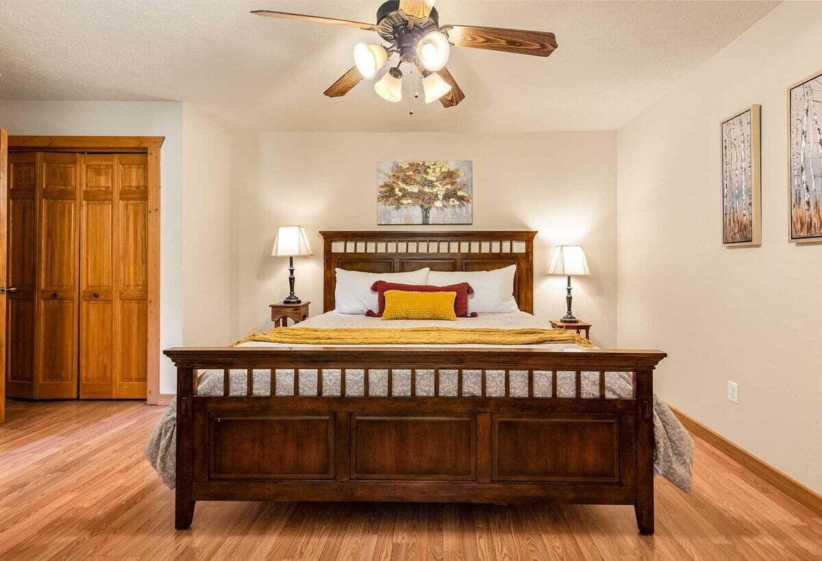 A bedroom with a wooden bed, two bedside lamps, a ceiling fan, and a door to the left.