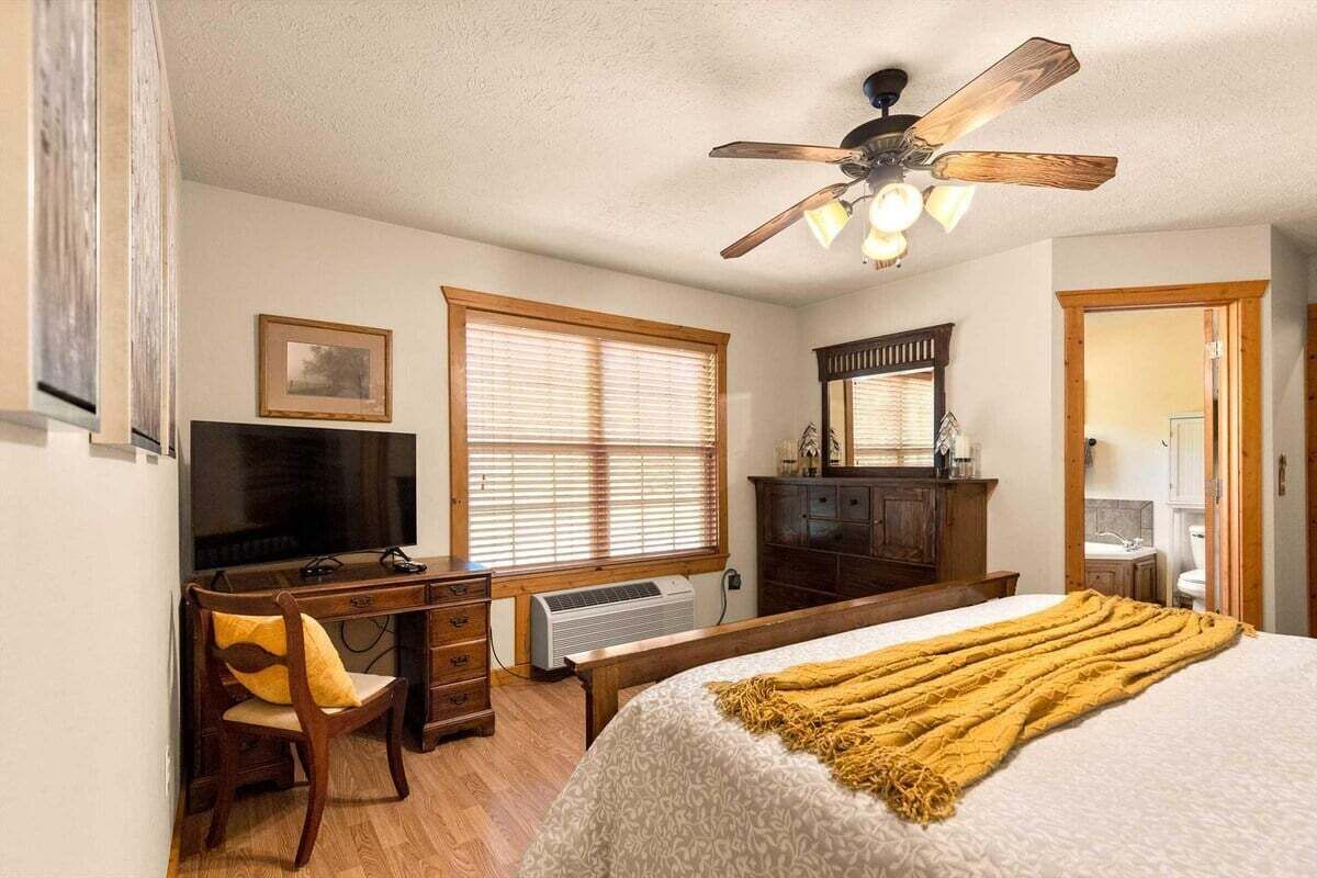 A bedroom with a bed, a desk, a chair, a ceiling fan, and a window with blinds.