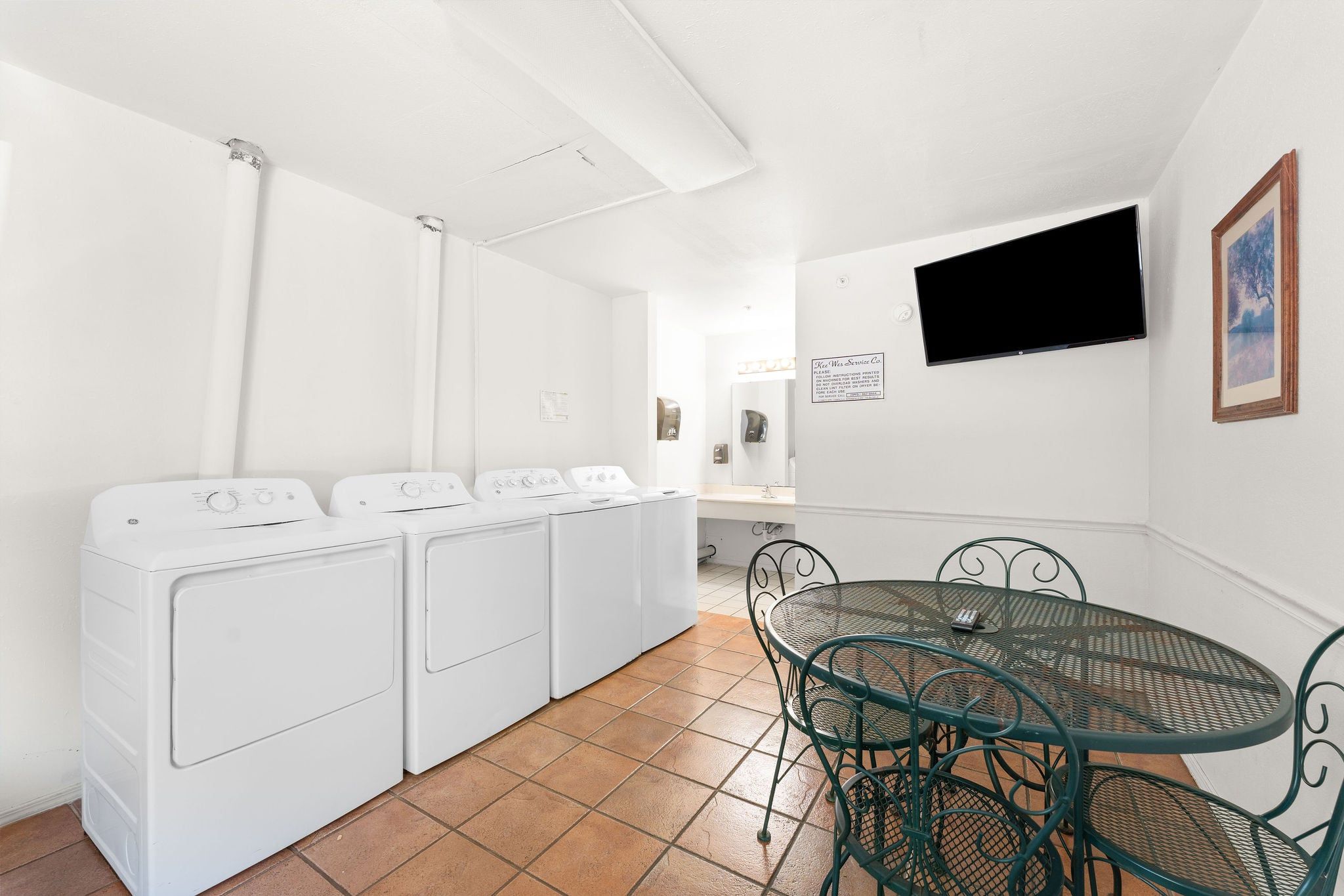 A laundry room with four white washing machines, a green metal table with chairs, and a mounted television on the wall.