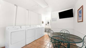 A laundry room with four white washing machines, a green metal table with chairs, and a mounted television on the wall.