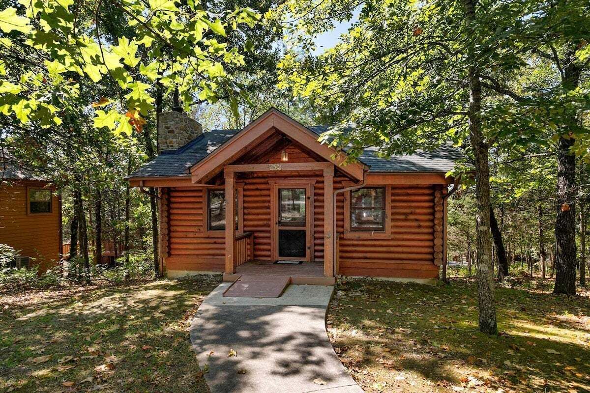 A log cabin with a front porch, a stone chimney, and a surrounding wooded area.
