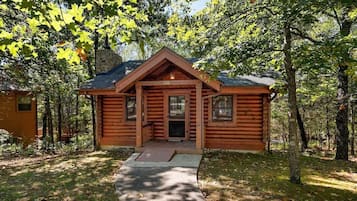 A log cabin with a front porch, a stone chimney, and a surrounding wooded area.