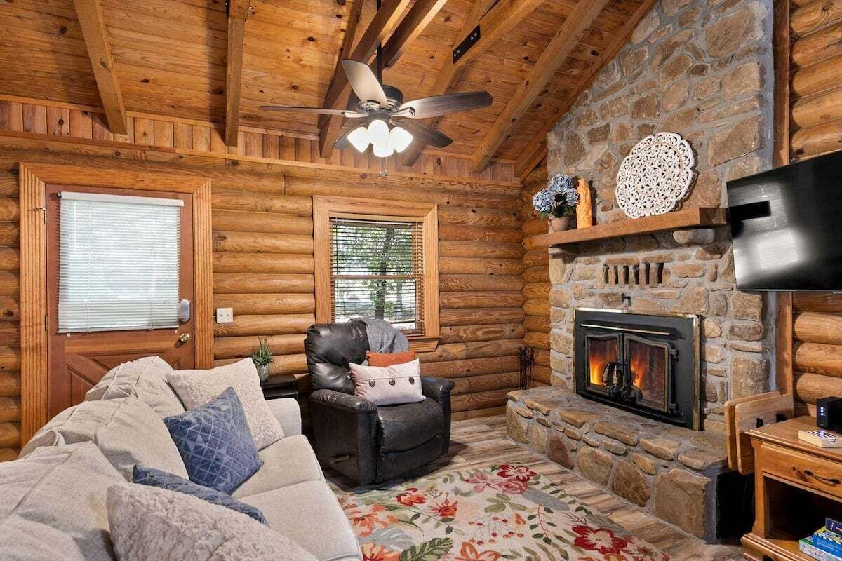 A log cabin interior with a fireplace, a leather chair, a sofa, and a ceiling fan.
