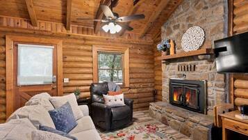 A log cabin interior with a fireplace, a leather chair, a sofa, and a ceiling fan.