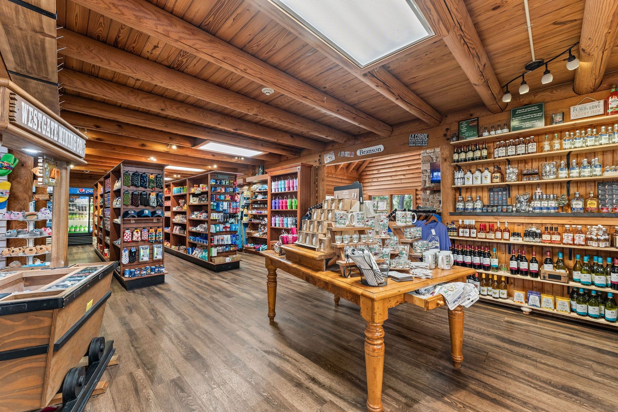 A wooden interior with a central table displaying various items, shelves stocked with bottles, and a 'Peace, Love, Country Music' sign.