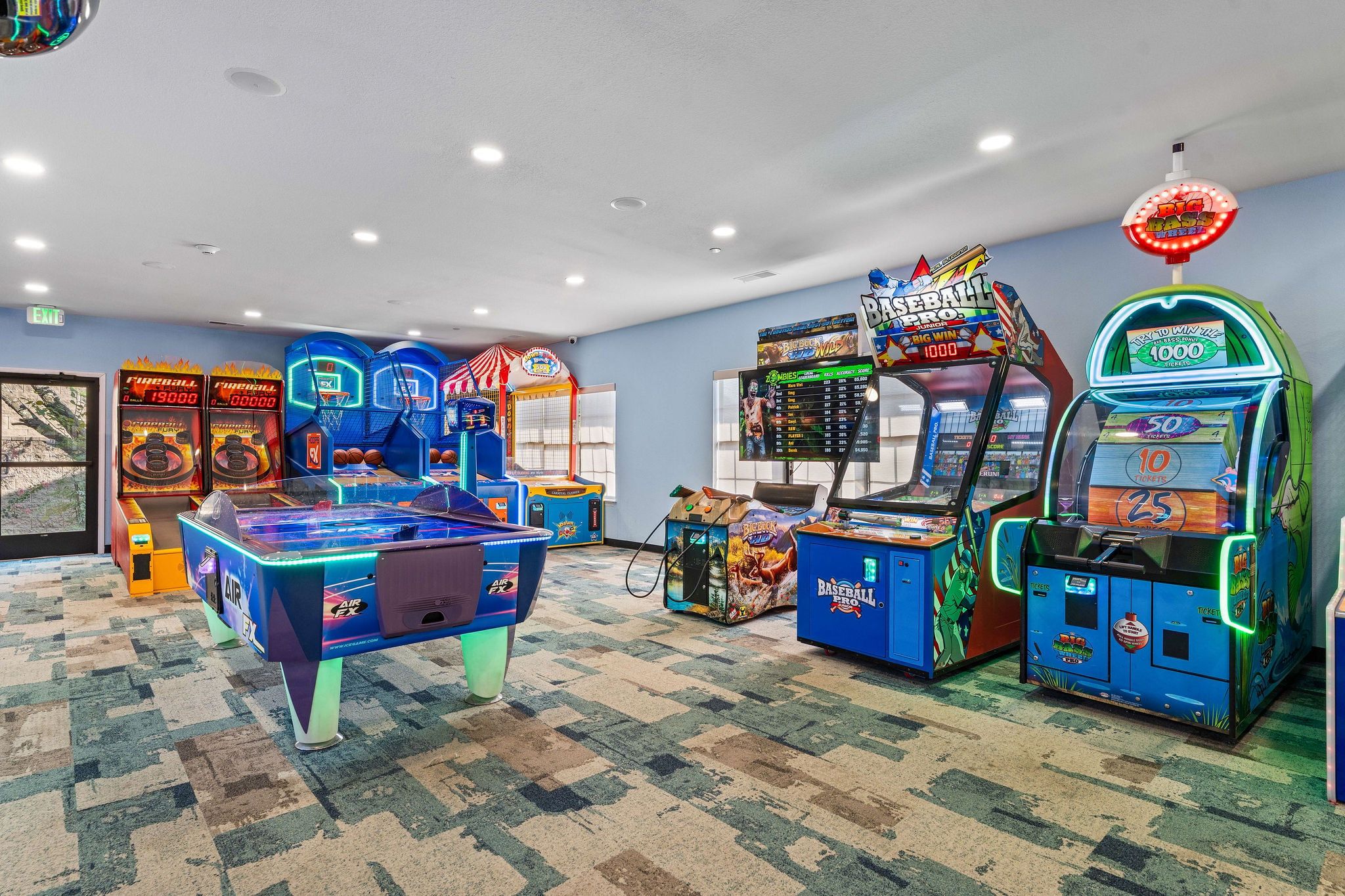 Habitación con máquinas de videojuegos arcade, incluyendo baloncesto y béisbol, piso alfombrado e iluminación empotrada en el techo.