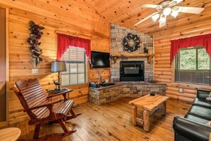 A wooden cabin interior with a fireplace, a ceiling fan, and a stone wall. - Luxury Log Cabin! King Bed, Full Kitchen, Fireplac (Branson)