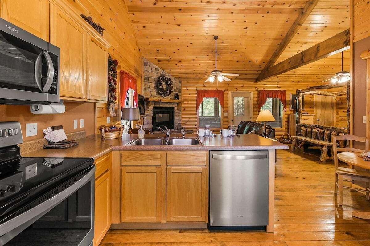 A cabin interior with wooden walls, a kitchen with a microwave, stove, and dishwasher, a fireplace, and a dining area with a bench and table.