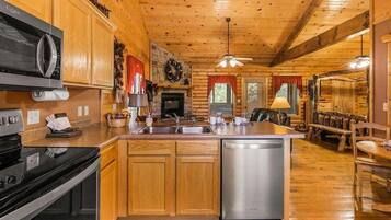 A cabin interior with wooden walls, a kitchen with a microwave, stove, and dishwasher, a fireplace, and a dining area with a bench and table.