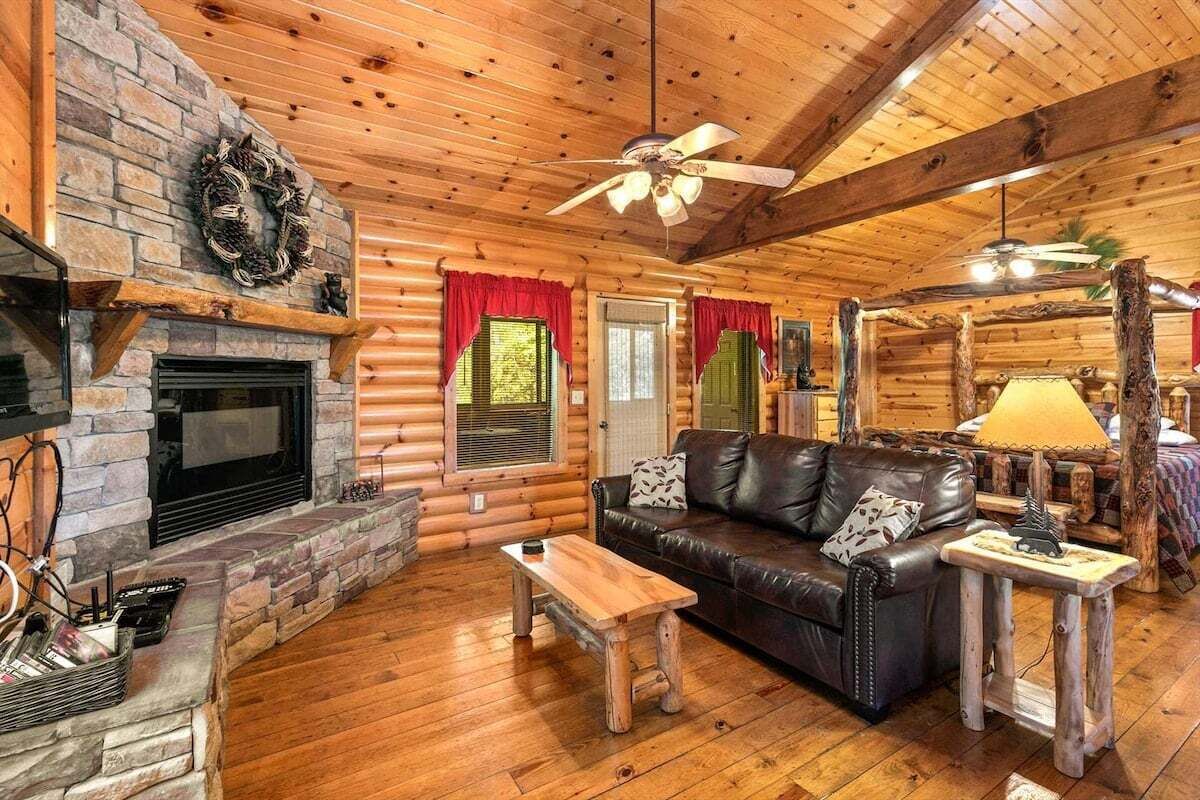 A log cabin interior with a fireplace, leather sofa, wooden coffee table, and a ceiling fan.