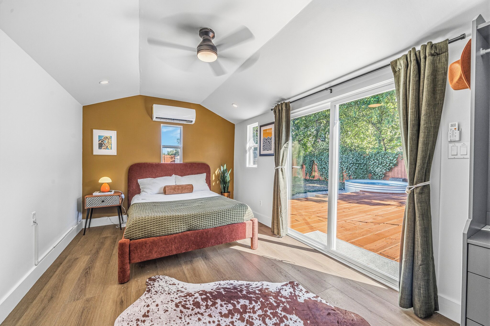 A bedroom with a bed, a ceiling fan, an air conditioning unit, a window with curtains, and a view of the outdoors.