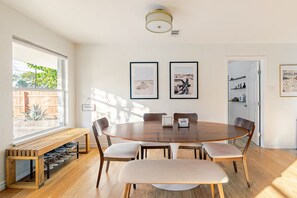 A dining area with a round wooden table, six chairs, a bench, and a window with a view of a wooden fence and greenery. - 4BR Austin Backyard Oasis/Cowboy Pool + Fire Pit / Near Zilker, SoCo & Downtown (Austin)