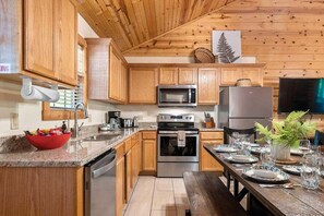 A kitchen with wooden cabinets, stainless steel appliances, a granite countertop, and a dining area set for four. - Resort Family Cabin_Incredible Amenities_Free Daily Attraction Tickets (Branson)
