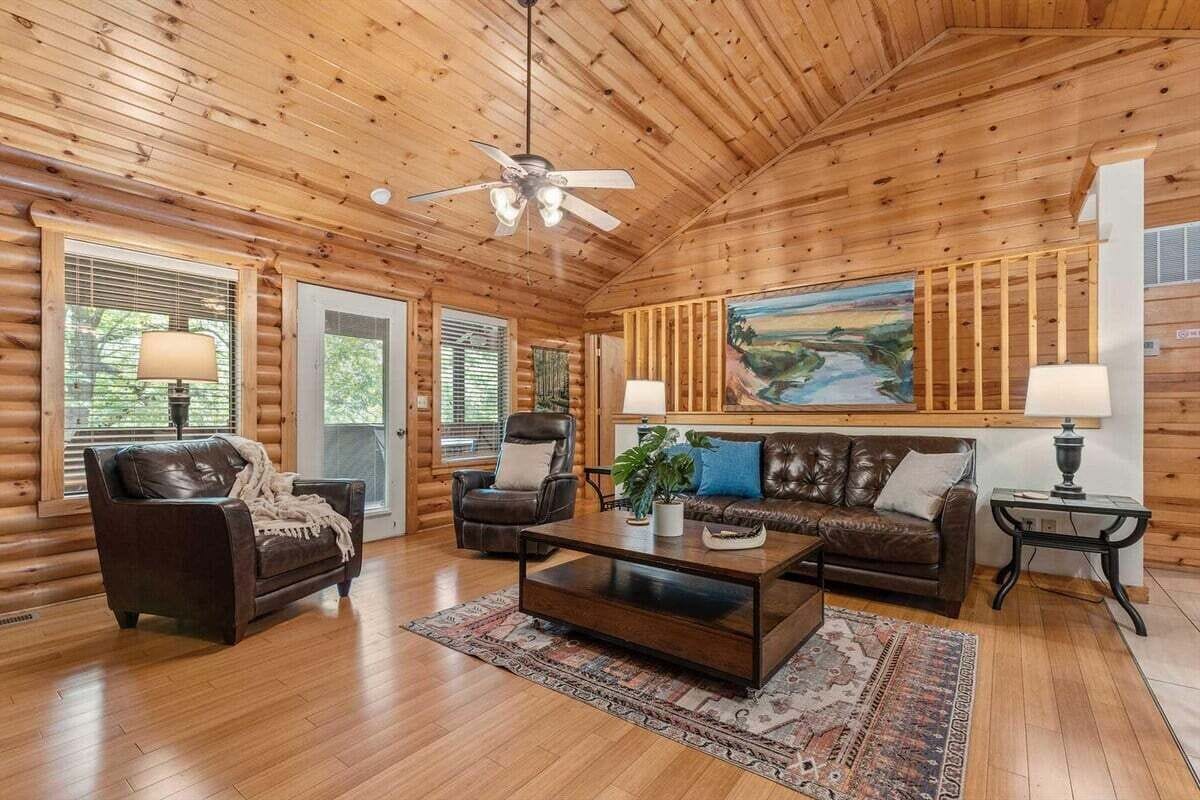 A log cabin interior with a wooden ceiling, a ceiling fan, a large painting, leather sofas, a coffee table, and two lamps.