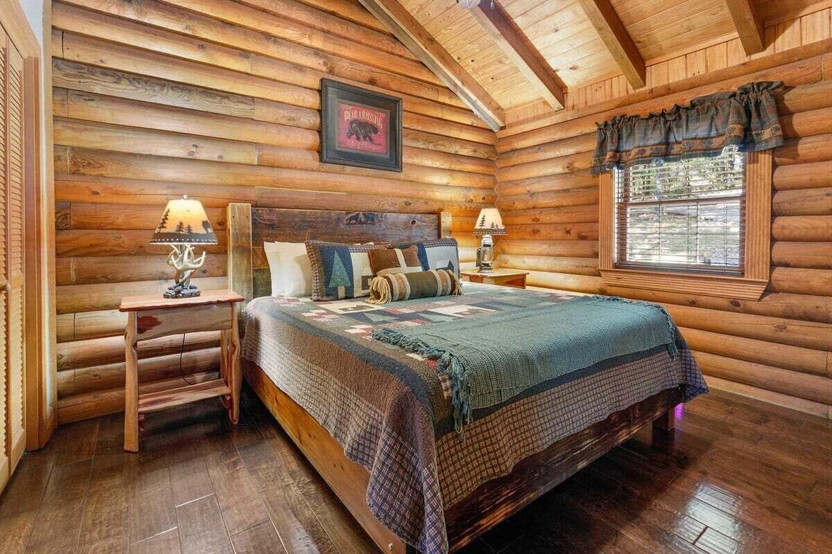 A log cabin bedroom with a bed, two nightstands, a window with curtains, and a framed picture on the wall.