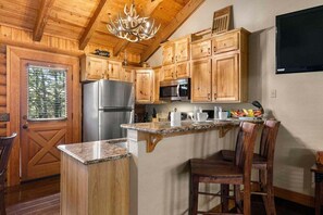 A kitchen with wooden cabinets, a stainless steel refrigerator, a microwave, and a granite countertop island with bar stools. - Incredible Double King Suite Cabin w Amazing Shared Amenities and Free Daily Attraction Tickets! (Branson)