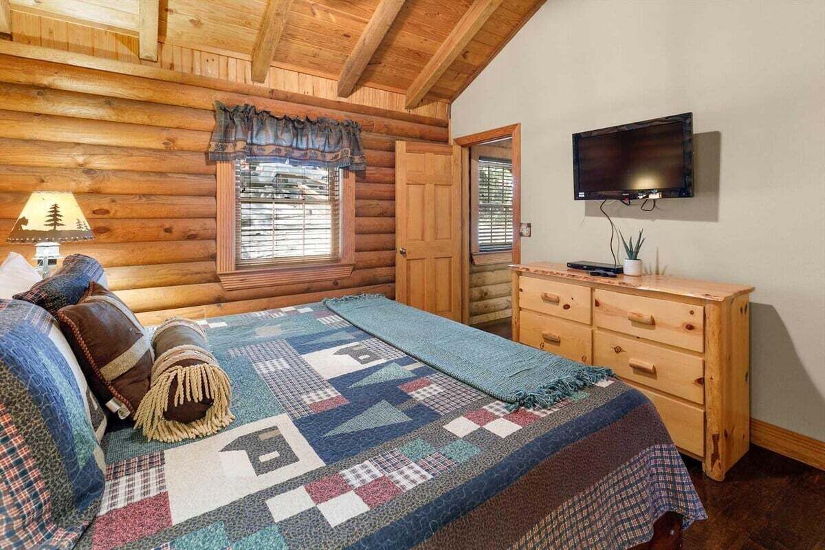 Une chambre située dans un chalet en rondins, avec un lit, une commode, un téléviseur et une fenêtre avec des stores.