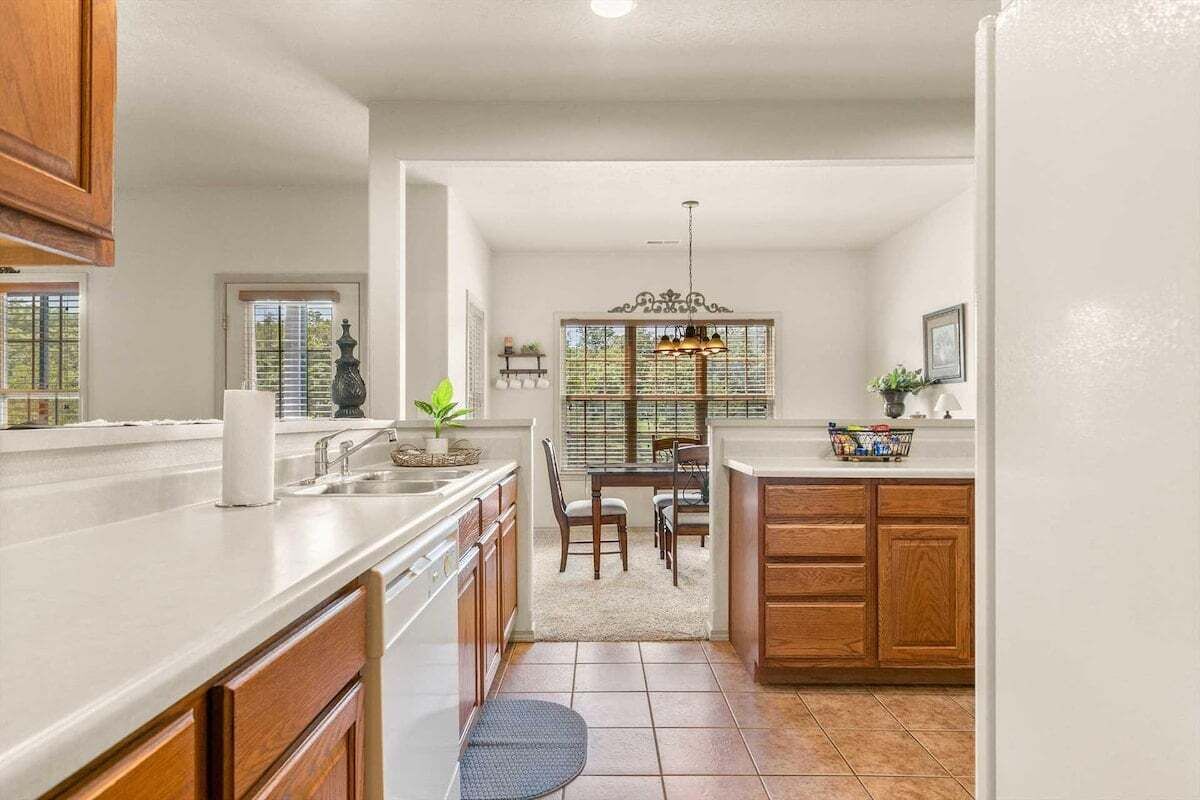 A kitchen with wooden cabinets, a white countertop, a dining area with a table and chairs, and a window with blinds.