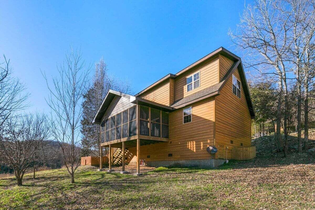A two-story wooden house with a large glass balcony, surrounded by trees and a grassy area.