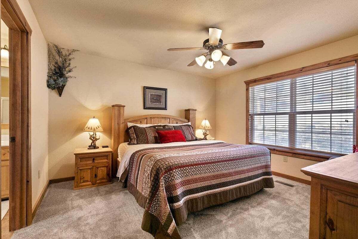 A bedroom with a bed, bedside lamps, a ceiling fan, a window with blinds, and a picture on the wall.