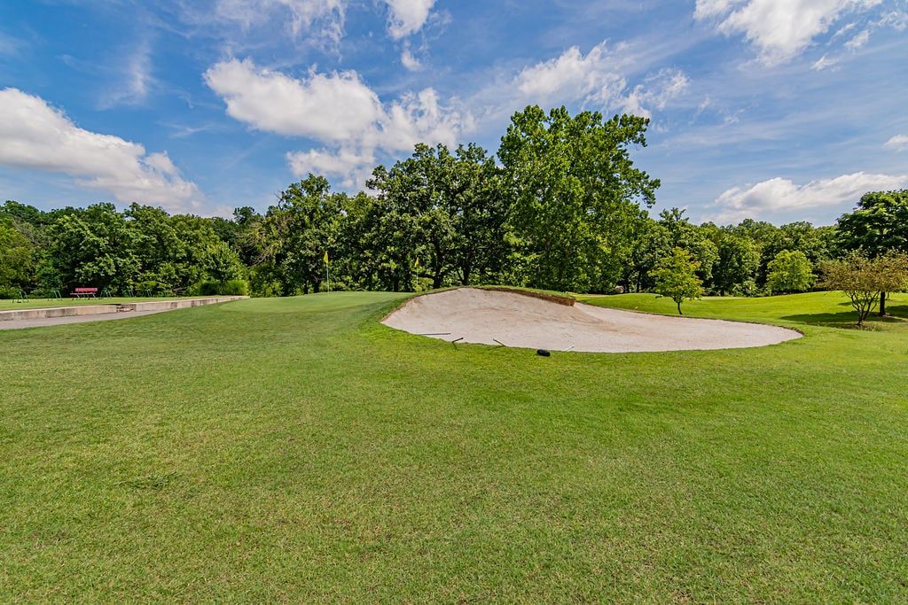 Een golfbaan met een zandbunker, goed onderhouden gras en een achtergrond van dichte bomen onder een lichtbewolkte lucht.