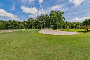 Campo de golf con un búnker de arena, césped bien cuidado y una densa arboleda al fondo bajo un cielo parcialmente nublado.