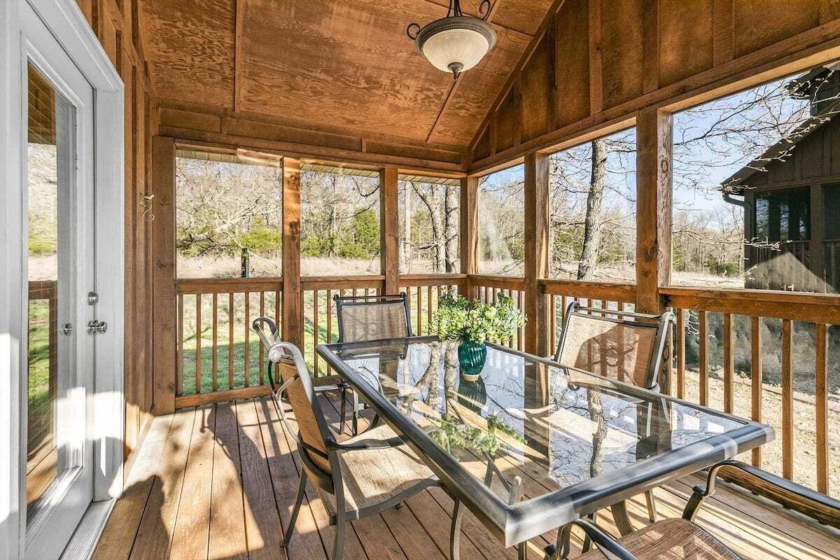 A covered outdoor area with a glass table and chairs, wooden deck, and a view of the surrounding landscape.