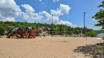 A sandy beach with a volleyball net, a children's play structure, and a backdrop of trees and buildings.
