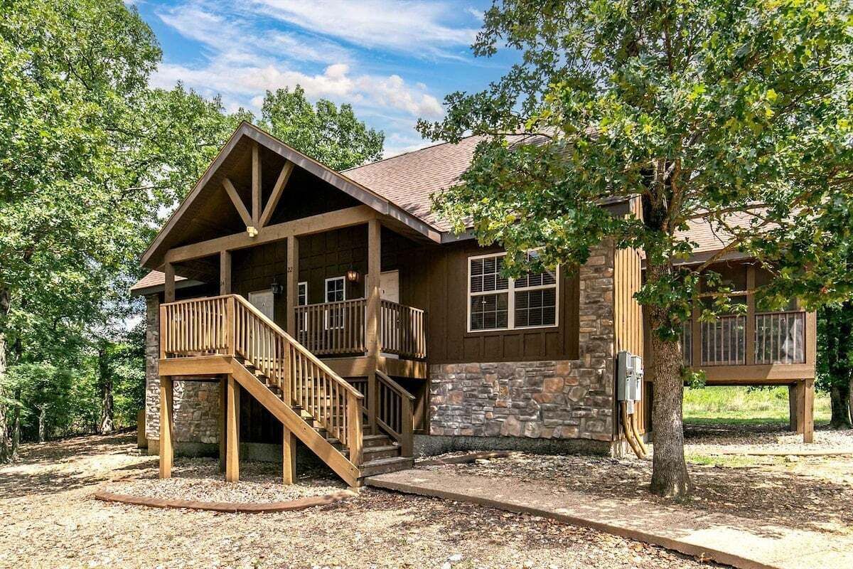 A two-story cabin with a stone exterior, a wooden deck, and a staircase leading to the upper level.