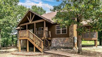 A two-story cabin with a stone exterior, a wooden deck, and a staircase leading to the upper level.