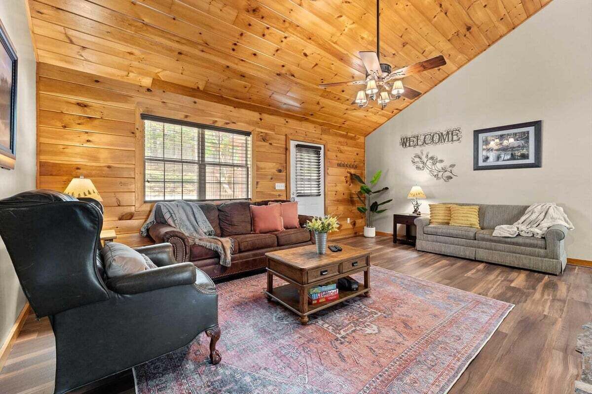 Un salon chaleureux avec un plafond en bois, un fauteuil en cuir noir, un canapé marron, un canapé gris et une table basse surmontée d’un tapis.