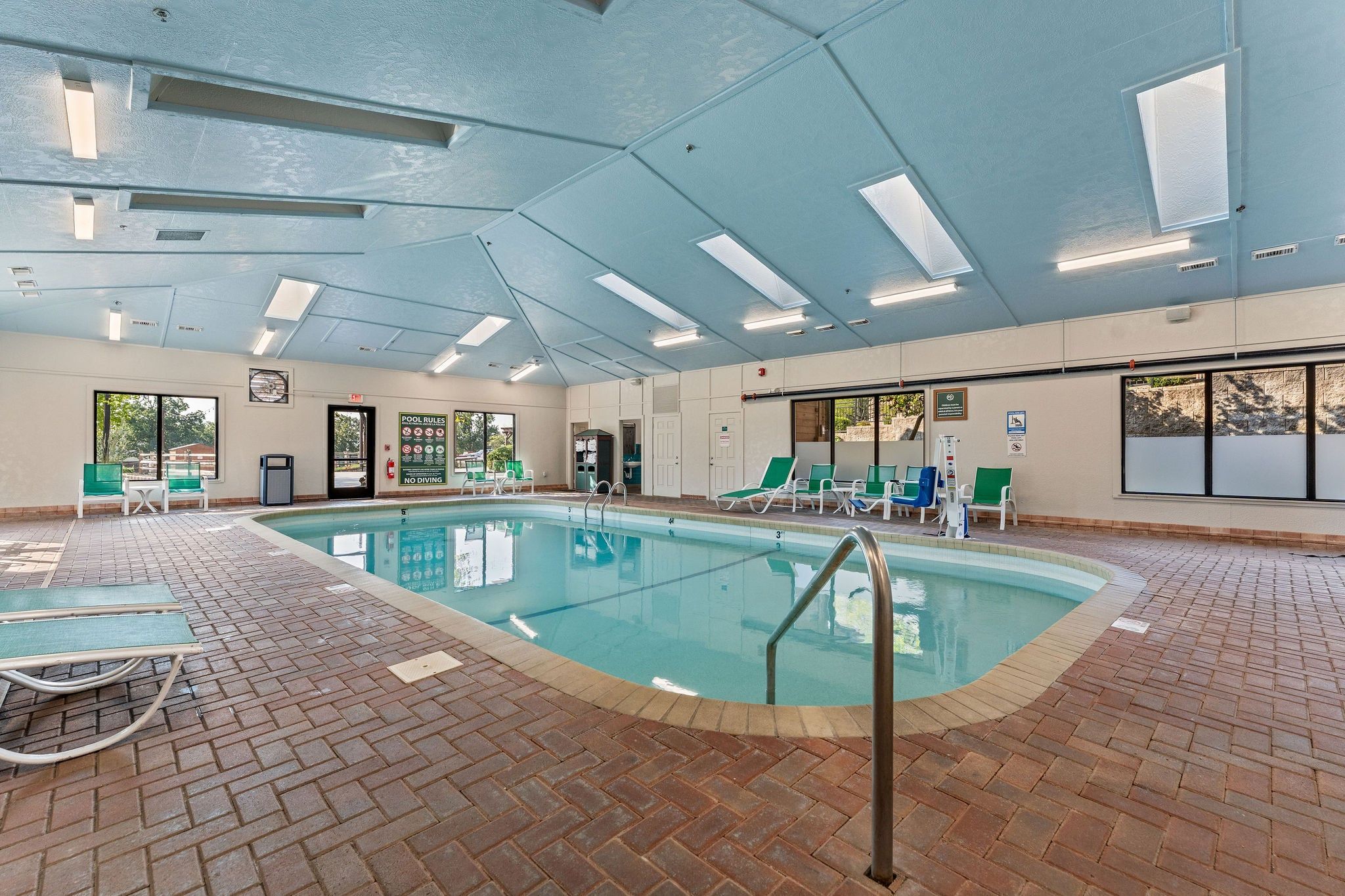 An indoor swimming pool with clear pool rules, a no diving sign, and a variety of lounge chairs around the pool area.